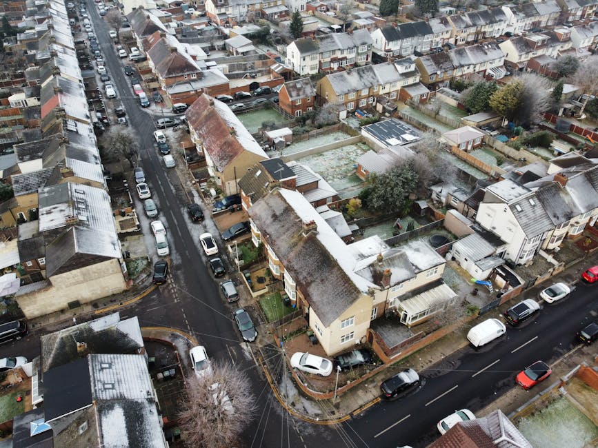An aerial view of a residential area showing narrow streets lined with parked cars and houses with small front gardens. The gardens feature various plants, trees, and lawn areas, some with snow or fro