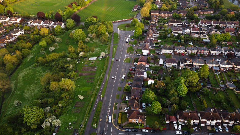 An aerial view of a residential area showing narrow streets lined with parked cars and houses with small front gardens. The gardens feature various plants, trees, and lawn areas, some with snow or fro