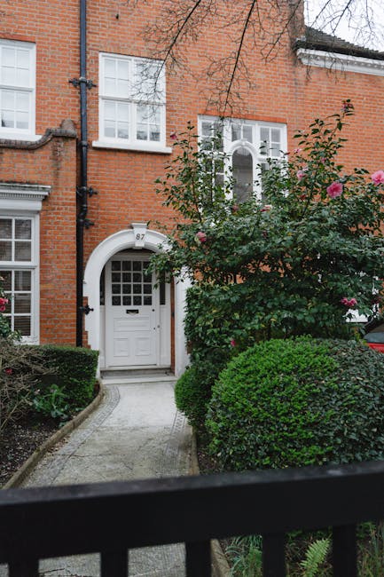 A front garden in Southfields featuring a paved pathway leading to a white front door set within a red-brick house. The garden is bordered by well-maintained, rounded green shrubs and a flowering shru